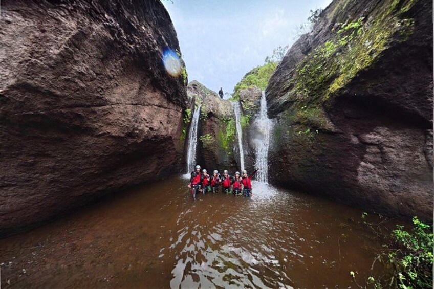 A group of people in red jackets standing in a river with a waterfall between two rock walls. 4