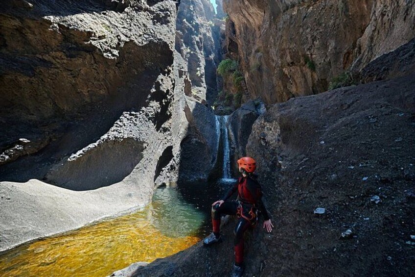 A person in a red helmet and black outfit sitting by a waterfall in a rocky canyon with a pool of water at the base. 3