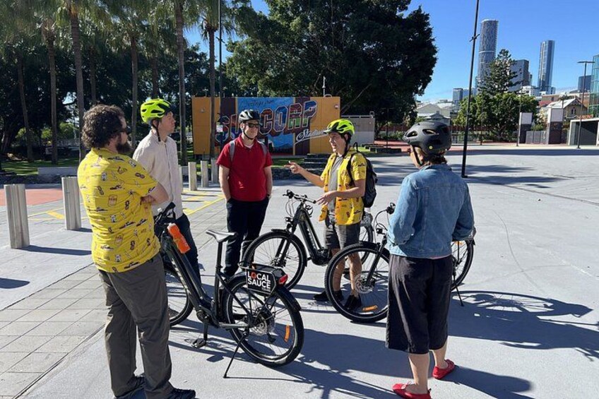 Un grupo de personas de pie en una acera con bicicletas, un letrero que dice 'SUGAR COCOA' y un paisaje urbano al fondo. 2