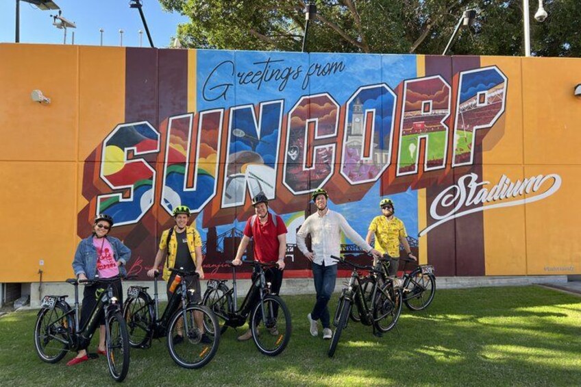 Un mural con el texto 'Greetings from SUNCORP Stadium' y un grupo de personas con bicicletas. 5