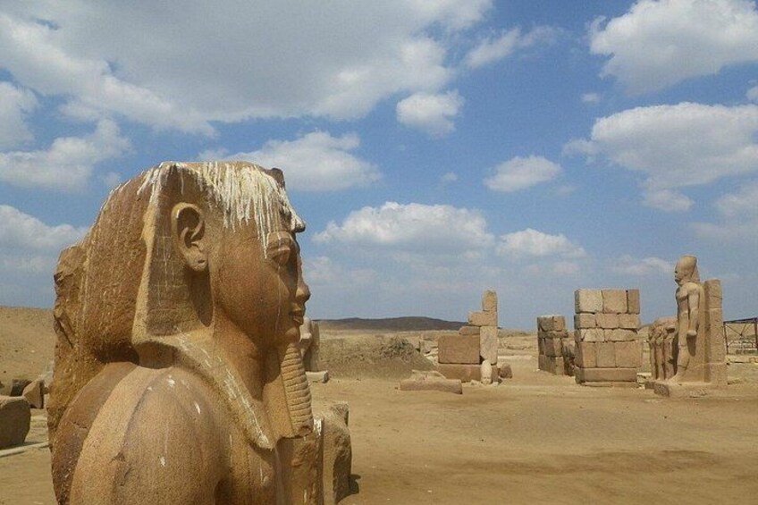 A large stone statue of a pharaoh in a desert landscape with scattered ruins in the background. 3