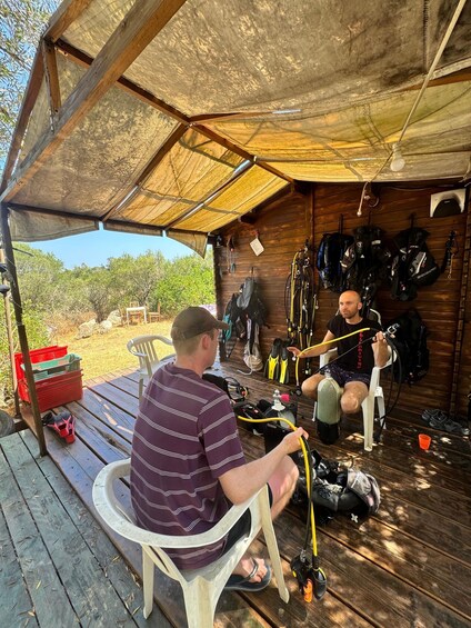 Two men sitting under a wooden structure with scuba diving equipment around them. 2