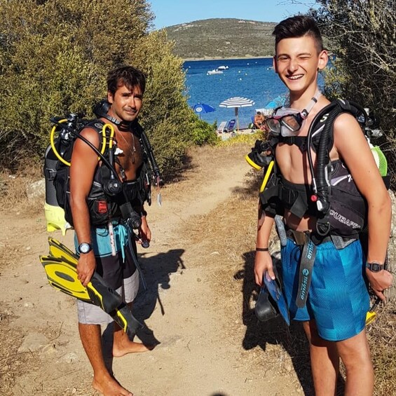 Two individuals with scuba diving gear, a sandy path, and a body of water with boats in the background. 3