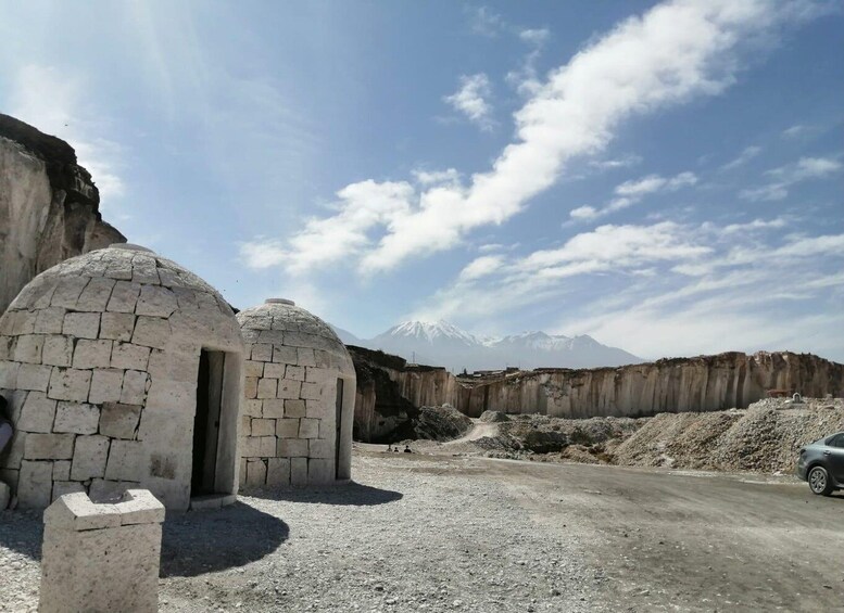 Un paisaje montañoso con dos construcciones de piedra que parecen yurtas, un camino de tierra y un coche aparcado cerca. 2