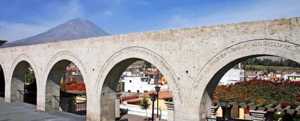 Un arco de piedra con inscripciones, una montaña al fondo y un paisaje urbano con edificios y vegetación. 5