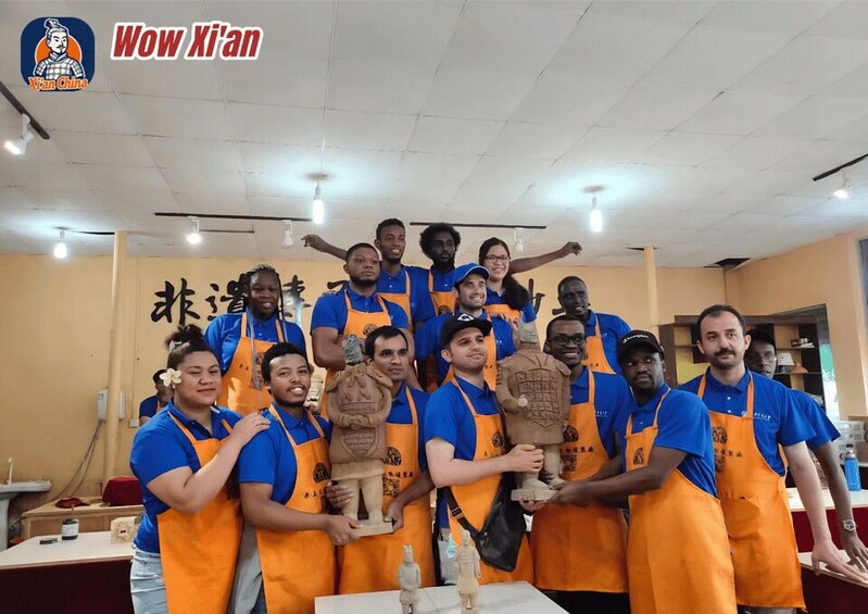 A group of people in a workshop environment, wearing blue shirts and orange aprons, holding various clay sculptures. 3