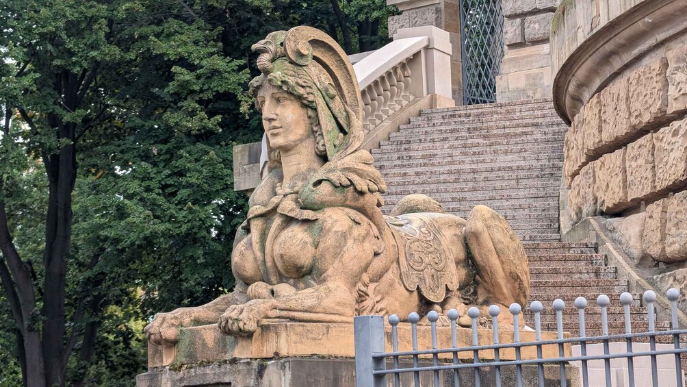 A stone sculpture of a reclining figure with a lion, set against a backdrop of trees and a stone staircase. 1
