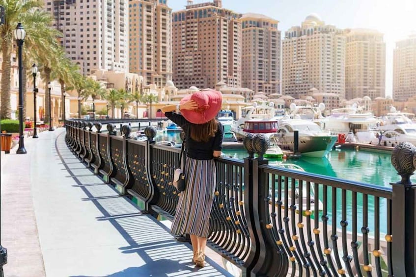 A woman standing on a waterfront promenade overlooking a marina with boats and tall buildings in the background. 2