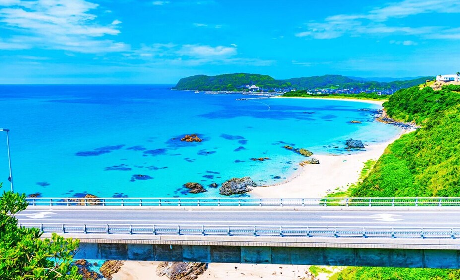 A coastal road with a beach, clear blue waters, and a hilly landscape with buildings. 2