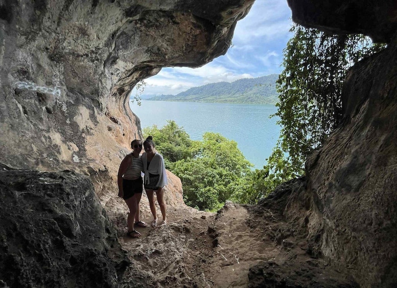 Two people standing at the entrance of a cave with a view of a lake and mountains in the background. 2