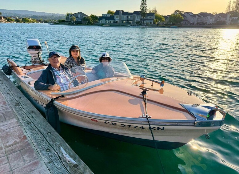 A small motorboat with three people on board, docked at a wooden pier with houses in the background. 1