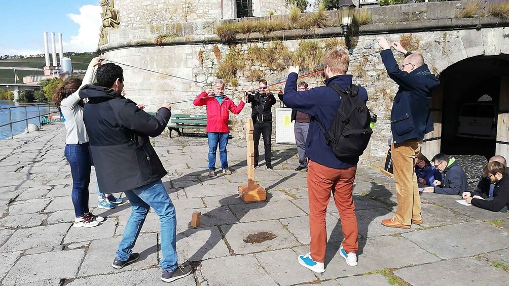 Folk deltager i en udendørs aktivitet, muligvis bueskydning, på en brostensbelagt plads tæt ved et vandareal og historiske bygninger. 5