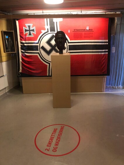 A room with a large red banner featuring a swastika and the text 'ET NAZISTISK NORGE'. In front of the banner is a podium with a bust on top. The floor has a red circle with text. 4