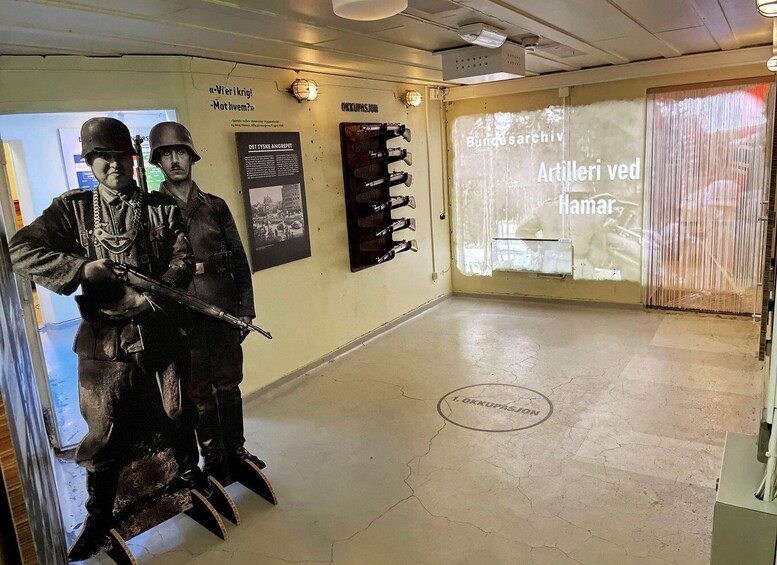A museum exhibit with a statue of soldiers, a projector displaying text, and a wall with mounted weapons. 5