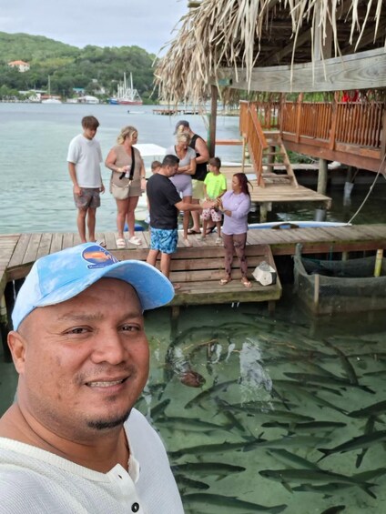 A man in a white shirt and blue cap standing on a wooden dock with a thatched roof, overlooking a body of water with several people and boats in the background. 4