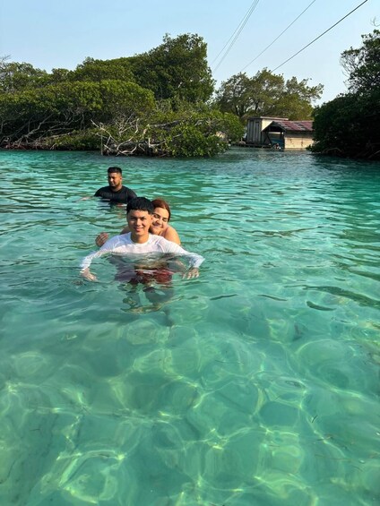 Two people swimming in clear turquoise water with a small house and dense greenery in the background. 2