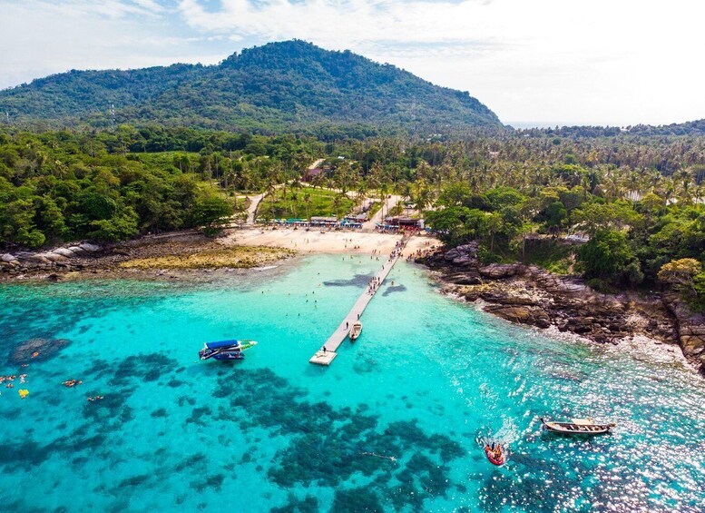 A coastal area with clear turquoise waters, a pier extending into the sea, and a mountainous backdrop. 1