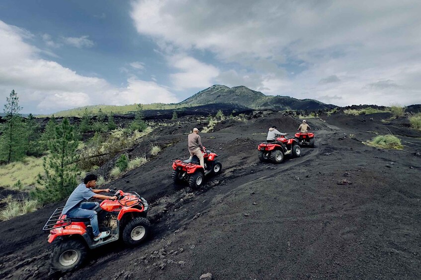 Three people riding red ATVs on a dirt road with a mountainous landscape in the background. 3