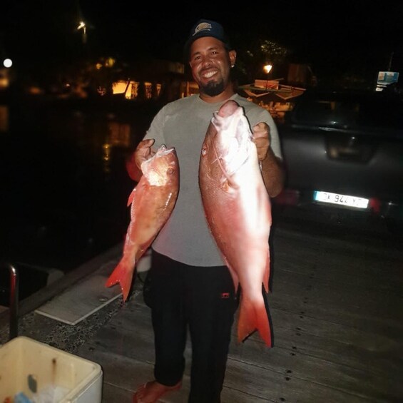 A person holding two large fish, standing outdoors at night with a vehicle and a building in the background. 5