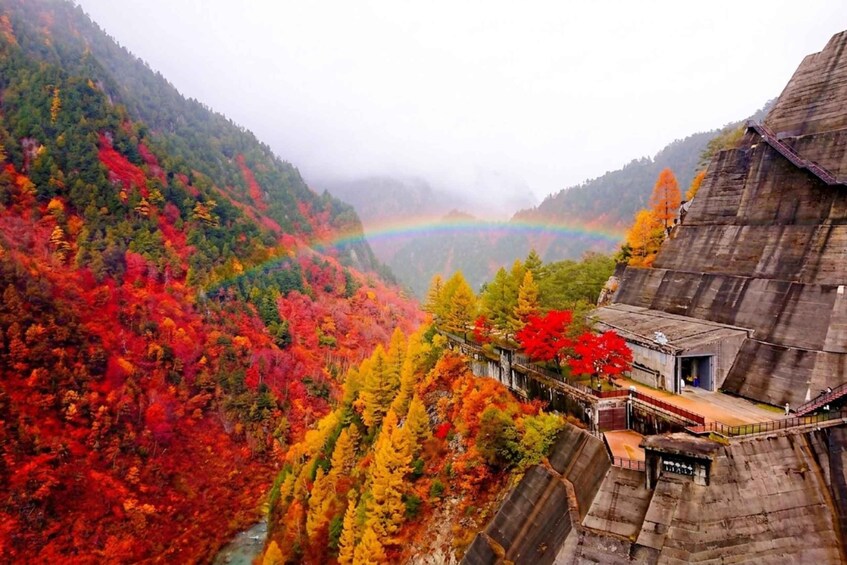 A mountainous landscape with a rainbow, autumn-colored trees, and a wooden structure. 1