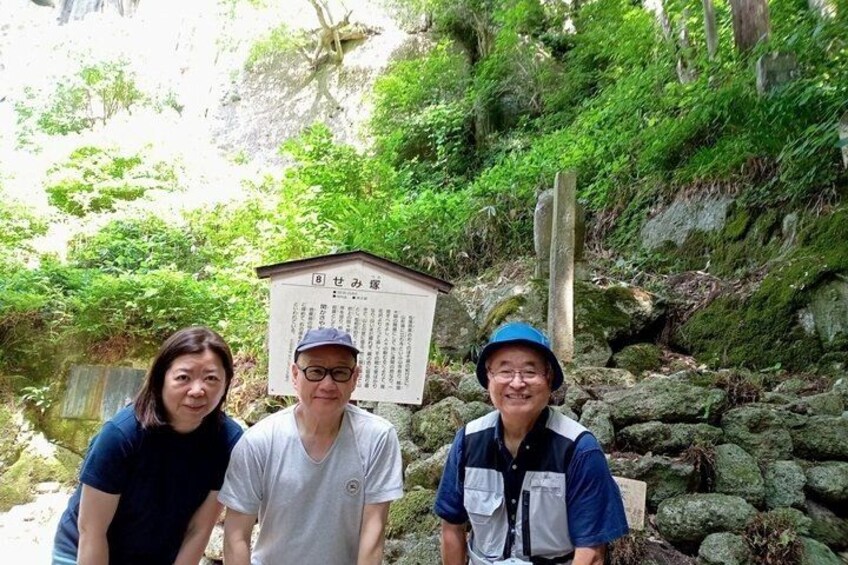 Tres personas frente a un cartel con texto japonés, rodeado de follaje y rocas. 2