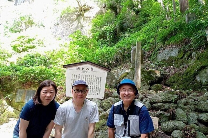 Three people standing in front of a sign with Japanese text, surrounded by greenery and rocks. 3