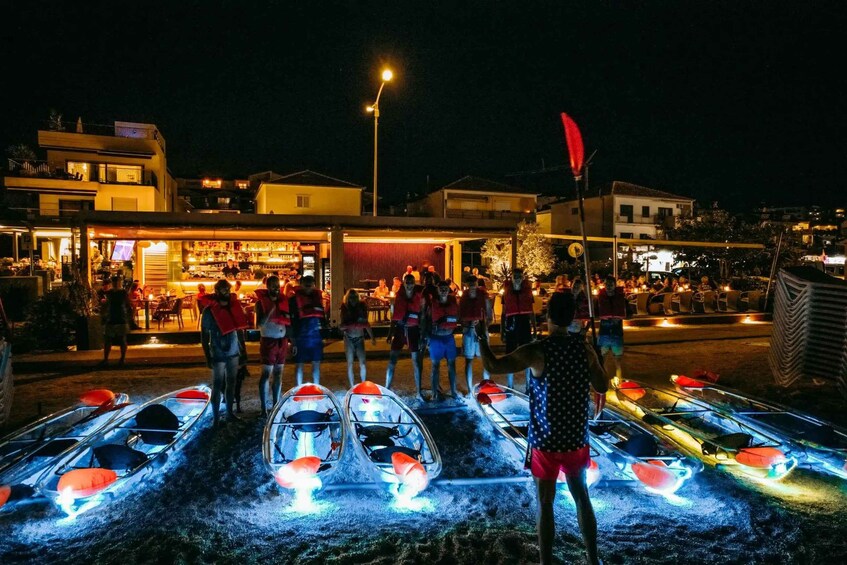 A nighttime scene with illuminated boats on a dock, people standing around, and buildings in the background. 3