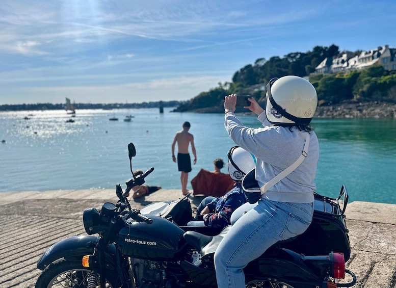 A person on a retro motorcycle by a waterfront with boats and houses in the background. 5