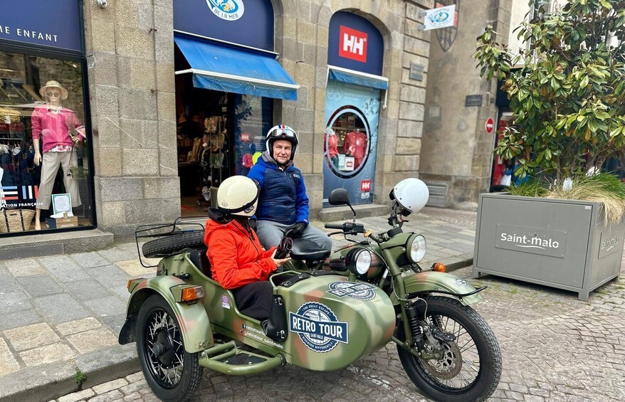A vintage motorcycle with a sidecar parked on a cobblestone street, a storefront with a blue awning, and a sign for Saint-Malo. 1