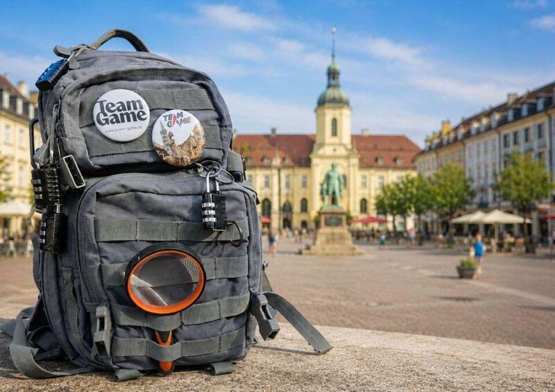 A large gray backpack with a circular orange and black object, a 'Team Game' sticker, and a keychain with a key. 1