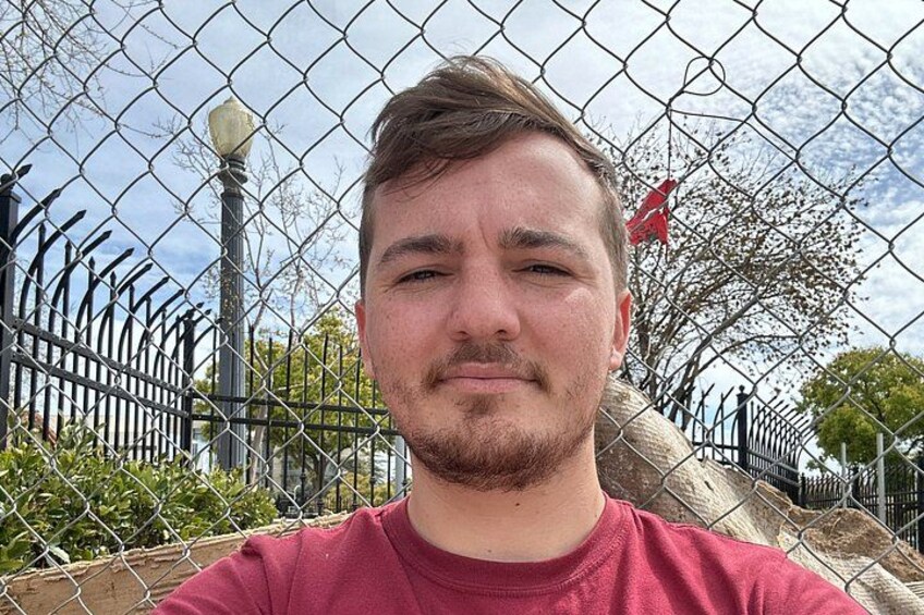 A person standing in front of a chain-link fence with a metal lamp post and trees in the background. 2