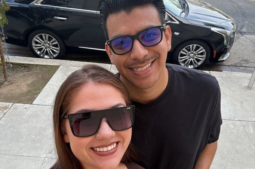 A man and a woman standing on a sidewalk with a black car parked in the background. 3