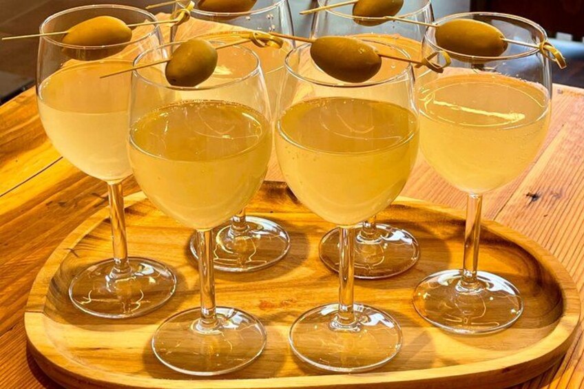 A wooden tray with several glasses of a yellow beverage, some with olive skewers. 3