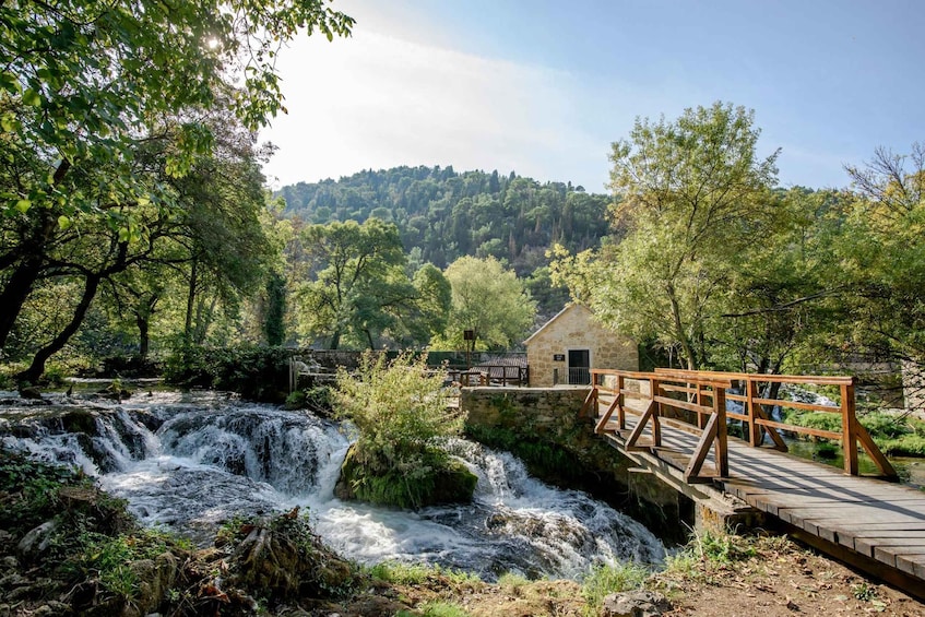 A wooden bridge over a waterfall, surrounded by lush greenery and a stone building. 2