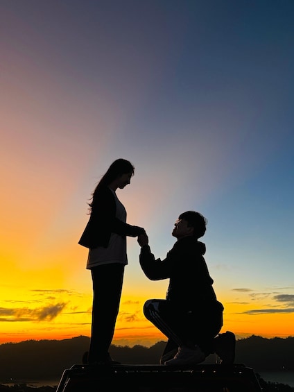 A silhouette of a person kneeling and holding hands with another person standing, against a sunset sky. 1