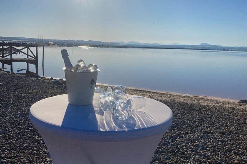 A table with a bucket of ice and glasses on a pebbled shore by a calm lake with distant mountains. 2