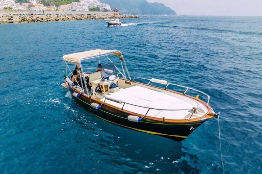 A small motorboat with a canopy, anchored in calm blue waters with a rocky shoreline and buildings in the background. 1