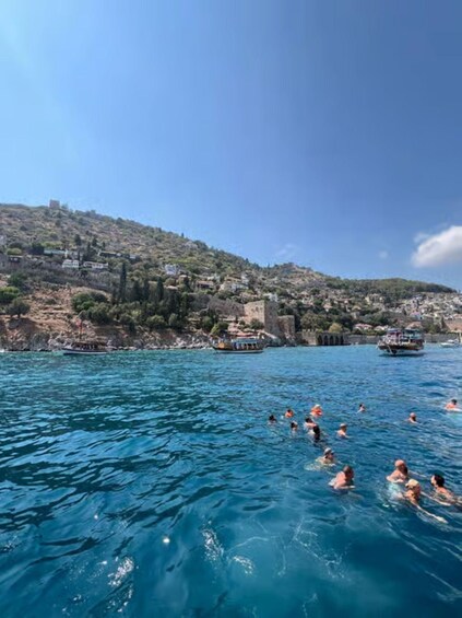 A coastal scene with people swimming in the water, boats, and a hillside with buildings. 3