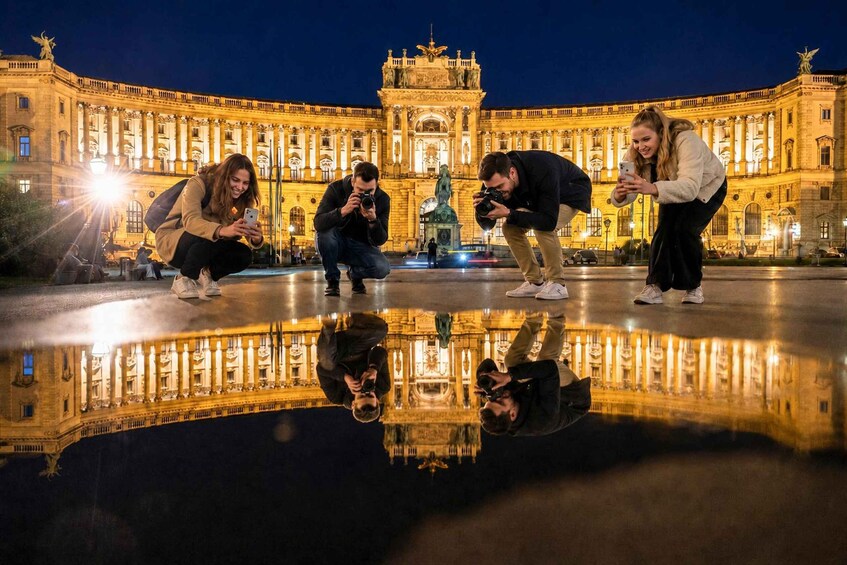 A group of people taking photos in front of a grand, illuminated building with a fountain in the center. 1