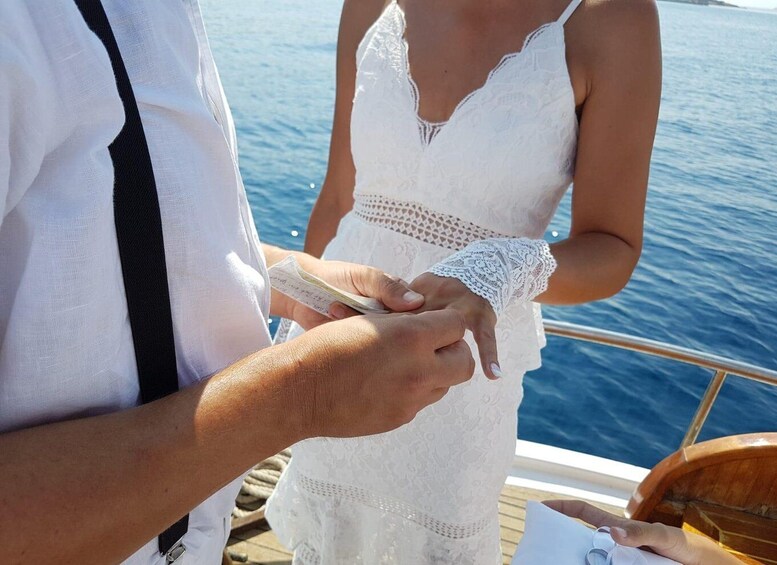 A person in a white dress holding a card, another person in a white shirt and black tie, and a wooden boat with a railing. 4