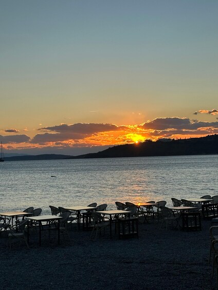 A sunset over a calm sea with chairs and tables on a pebbled beach. 2
