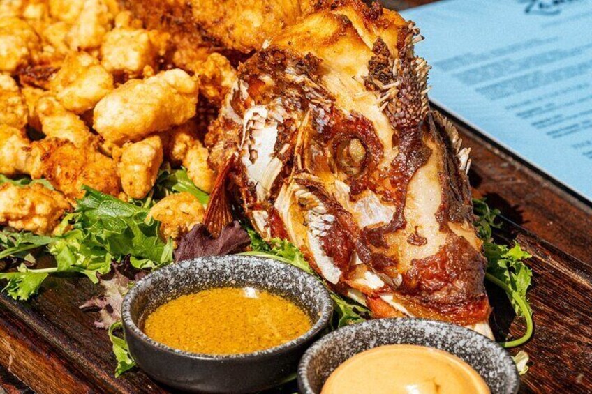 A whole grilled fish with crispy skin, accompanied by fried items and two small bowls of dipping sauces. 5