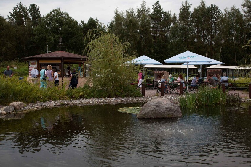 A lakeside area with people dining under umbrellas, a small pavilion, and a pond with a large rock in the center. 2