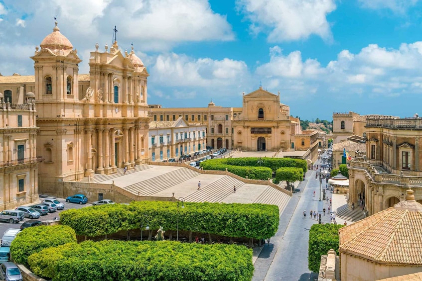 A historic plaza with ornate buildings, a central fountain, and neatly trimmed hedges. 1