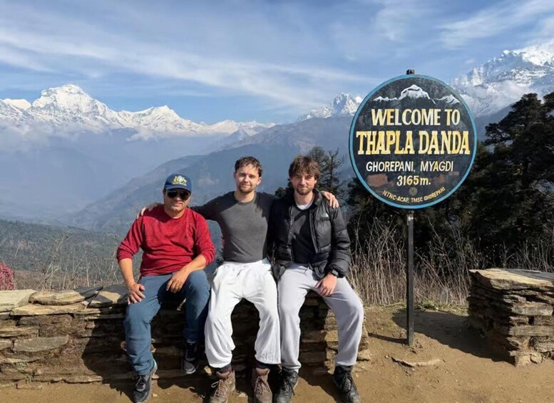 Three people sitting on a stone wall with a sign in the background welcoming visitors to Thapala Danda, Ghorepani, Myagdi, at 3165m elevation. 3