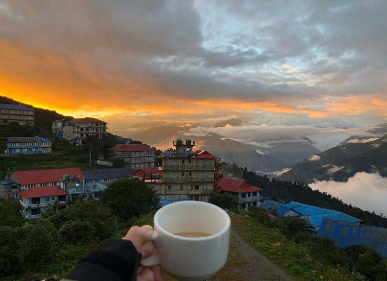 A person holding a cup, overlooking a mountainous landscape with houses and a sunset. 2