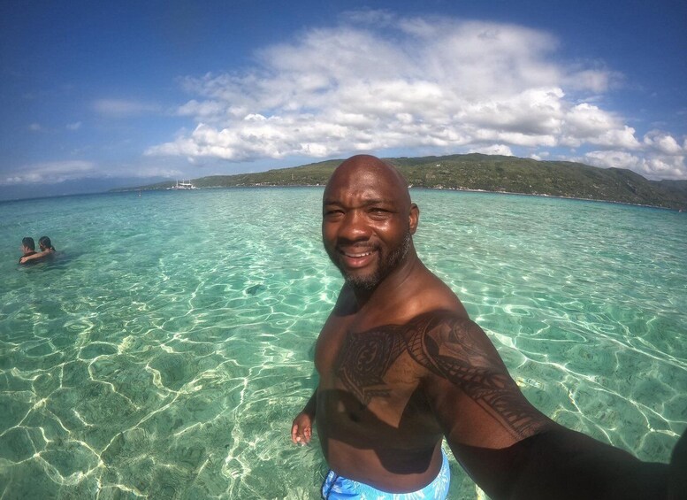 A man in a tropical setting with clear turquoise water, a distant boat, and a hilly landscape under a partly cloudy sky. 2