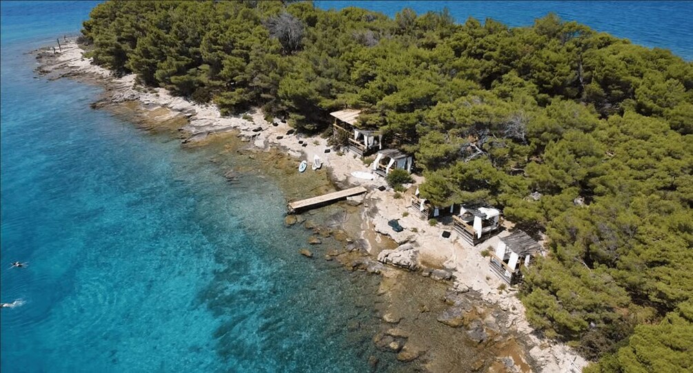 A coastal area with clear blue waters, a rocky shoreline, dense green trees, and a small wooden pier. 2