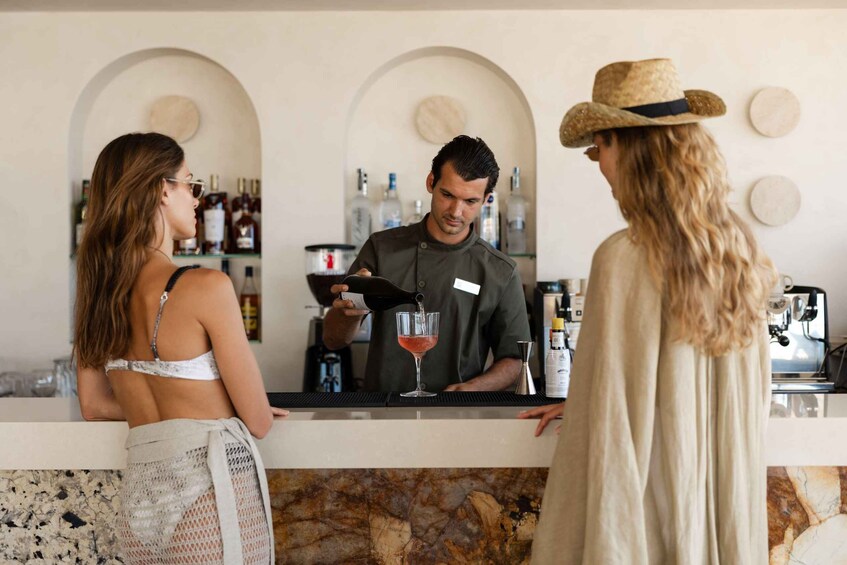 A bar setting with a bartender preparing a drink, a woman in a straw hat, and a woman in a white top. 3