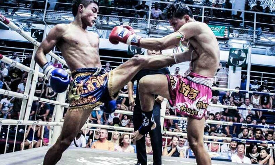 Two fighters in a boxing ring, one delivering a kick, with spectators in the background. 2